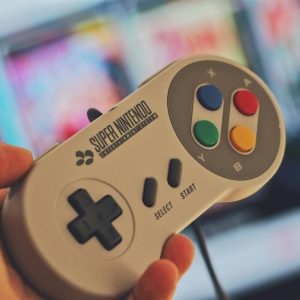 Hand Holding A Classic Super Nintendo Game Controller Indoors With A Blurred Screen In The Background.