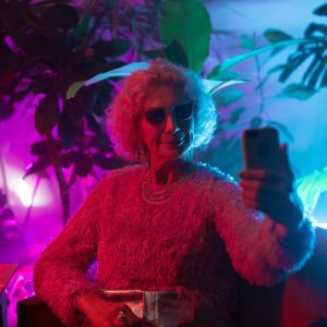 Chic Senior Lady With Sunglasses Takes A Selfie In Vibrant Neon Lighting With Indoor Plants.
