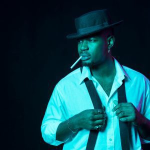 Confident Man In Black Hat And Suspenders With A Cigarette, Exuding Style Against A Dark Backdrop.