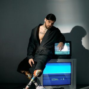 Bearded Man In Studio Sitting On Vintage CRT TV, Showcasing Technology Integration And Modern Style.
