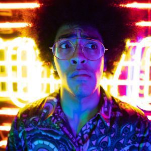 A Colorful Portrait Of A Young Man With Curly Hair And Glasses In Dramatic Neon Lighting.