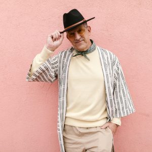 Fashionable Senior Man In A Hat And Scarf Poses Stylishly Against A Pink Wall.