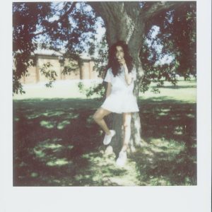 A Vintage Style Photograph Of A Woman In A White Dress Leaning Against A Tree Outdoors.
