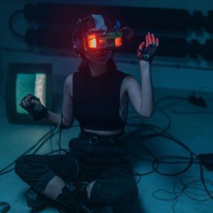 A Woman Wearing A VR Headset Sits On The Floor Surrounded By Retro Tech In A Dark Room.