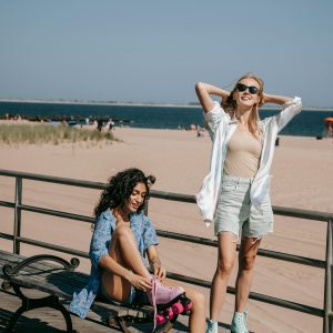 Two Women Enjoying A Sunny Day Roller Skating By The Beach On A Wooden Boardwalk.
