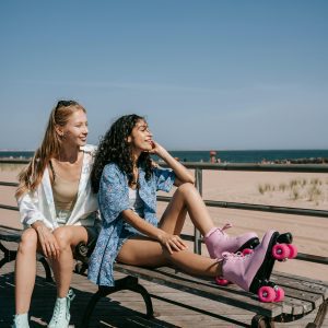 Two Young Women In Roller Skates Enjoying A Sunny Day At The Beach On A Bench.