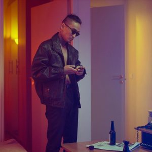 A Man In A Vintage Leather Jacket Using His Smartphone In A Warm Lit Room With An Old TV And Lamp.
