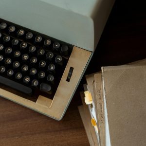 A Classic Vintage Typewriter Alongside Stacked Brown Paper Documents On A Wooden Desk.
