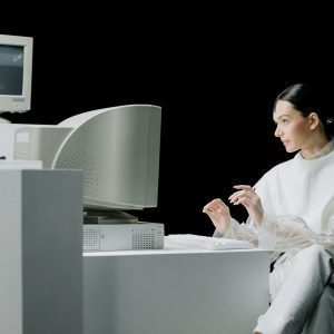 A Woman In A Futuristic Outfit Interacts With A Vintage Computer In A Minimalist Environment.