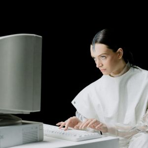 Woman In Futuristic Attire Engaging With A Vintage Computer In A Studio Setting.