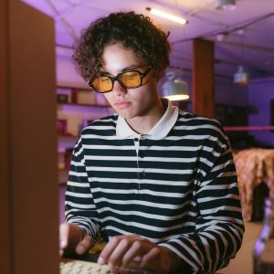 A Young Man Wearing Sunglasses Types On A Retro Computer Keyboard In A Cozy Indoor Workspace.
