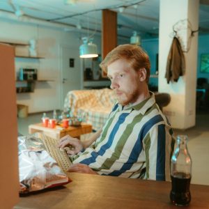 Man In Striped Shirt Typing On Keyboard In Vintage 80s Inspired Room With Snacks And VHS Tapes.
