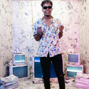 Young Man In A Vibrant Shirt And Sunglasses In A Retro Themed Room Decorated With Outdated TVs And Newspapers.