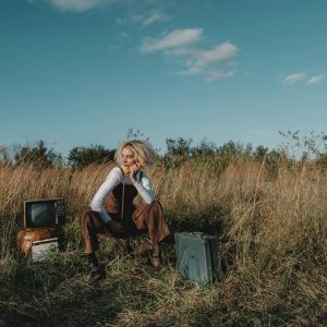 Stylish Woman Posing With Vintage TV And Suitcase In A Sunny Grassland Setting.