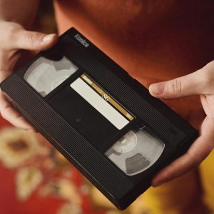 A Person Holds A Vintage VHS Tape Against A Warm, Patterned Background, Evoking Nostalgia.