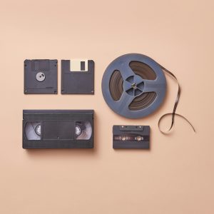 A Collection Of Vintage Media Storage Formats Including VHS, Cassette, And Floppy Disks On A Beige Background.