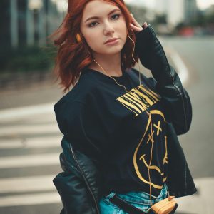Fashionable Young Woman With Red Hair In Leather Jacket And Headphones On City Street.