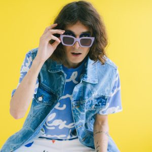 Portrait Of A Stylish Adult Wearing A Denim Jacket Against A Vibrant Yellow Background.