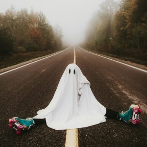A Whimsical Ghost In A Sheet Costume Sits On A Foggy Road With Roller Skates.