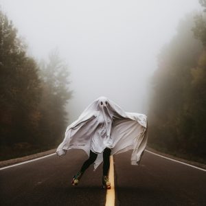 A Person In A Ghost Costume Roller Skates Down A Foggy Forest Road During Halloween.