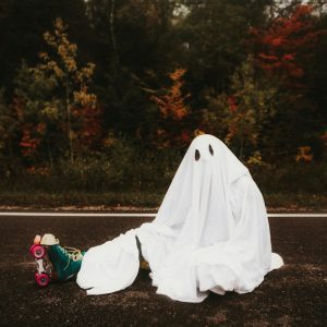 A Person In A Ghost Costume Sits On A Forest Road With Roller Skates, Surrounded By Autumn Foliage.
