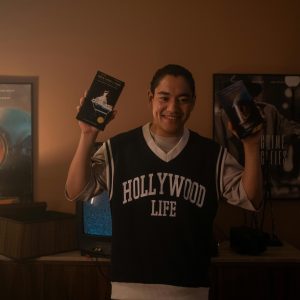 Smiling Man Holding Vintage VHS Tapes, Standing In A Warmly Lit Room With Classic Movie Posters.