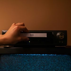 A Hand Inserting A VHS Tape Labeled 'SHINE' Into A Vintage Player. Nostalgic Home Movie Setup.