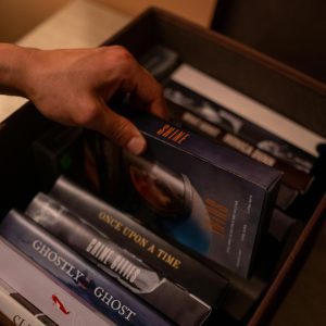 A Hand Selecting A VHS Tape From A Box Of Various Nostalgic Movie Covers Indoors.