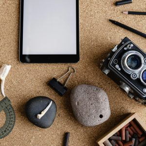 A Flatlay Of Vintage Camera, Tablet, Pencils, Stones, And Art Supplies On A Cork Desk.